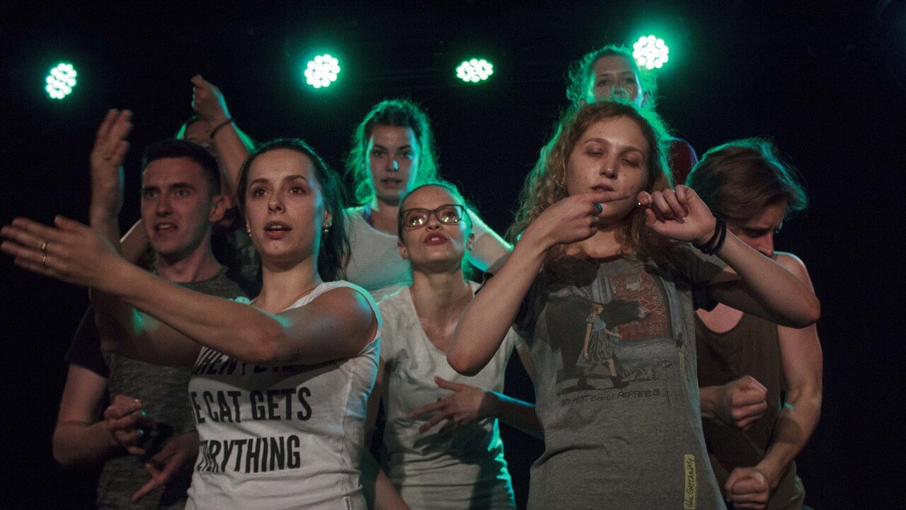 osiem osób stoi w grupie, wśród nich sześć kobiet i dwóch mężczyzn, wykonują dłońmi różne gesty, wyraz twarzy sugeruje, że coś mówią, z tyłu pada zielone światło, zdjęcie z akcji w sieci teatru gdynia główna
