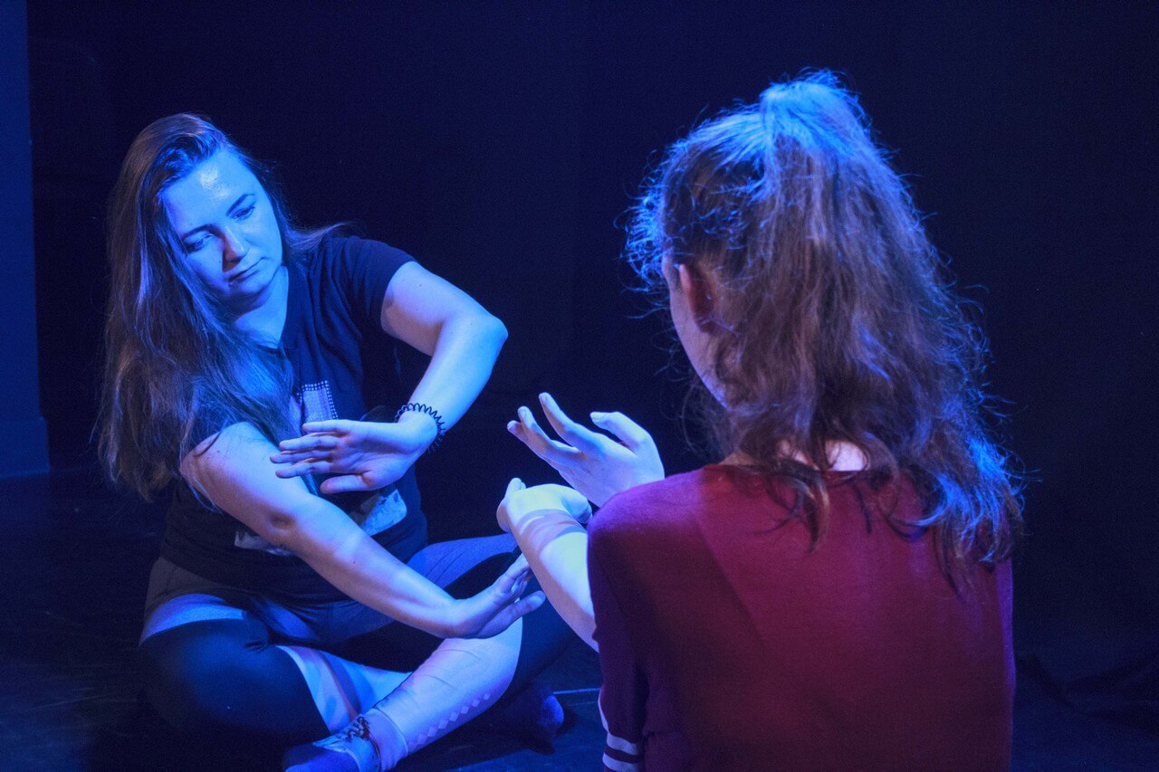 na ziemi siedzą dwie dziewczyny, wyciągają przed siebie dłonie w skomlikowanych gestach, pada niebieskie światło, zdjęcie z akcji W SIECI - miejska akcja Teatru Gdynia Główna w przestrzeni Dworca PKP