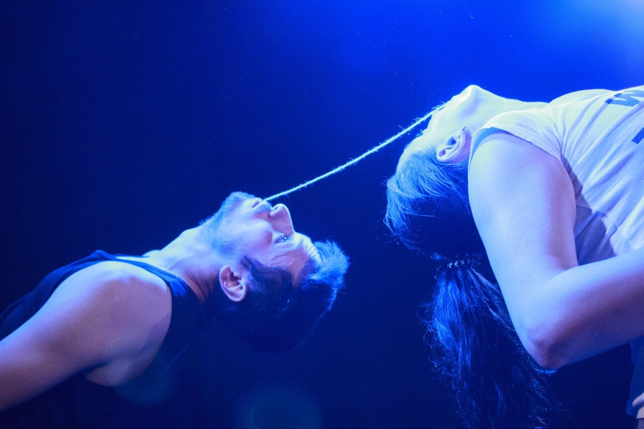 mężczyzna i kobieta odwróceni do siebie plecami odchylają się do tyłu, trzymają w ustach inne końce tego samego kawałka sznurka, pada niebieskie światło, zdjęcie z akcji W SIECI - miejska akcja Teatru Gdynia Główna w przestrzeni Dworca PKP
