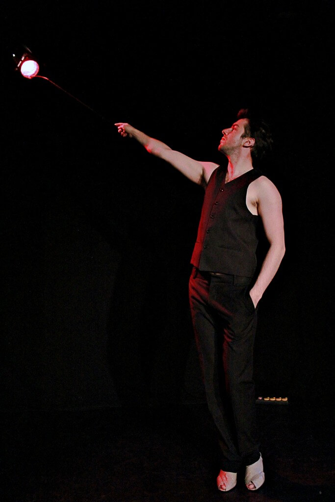 aktor o ciemnych włosach, ubrany w czarny strój, stoi z ręką wyciągniętą w lewą stronę zdjęcia, w kierunku reflektora