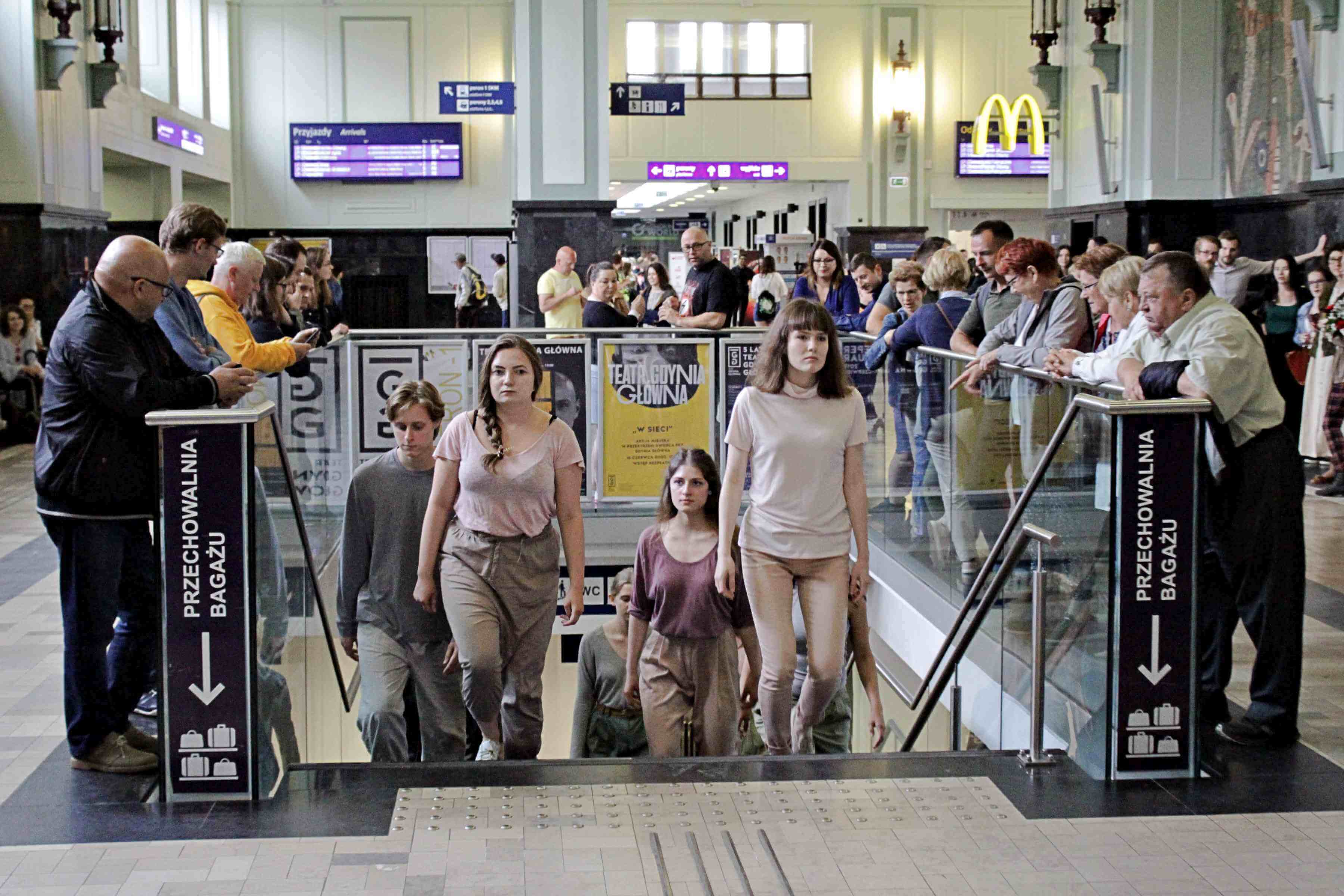 w przestrzeni dworca, po schodach prowadzących do teatru wychodzą z niego ustawieni parami młodzi ludzie, na przedzie idą dwie kobiety, wszyscy ubrani są w jasne kolory, odcienie szarego i różu, dookoła widać przyglądających się przechodniów