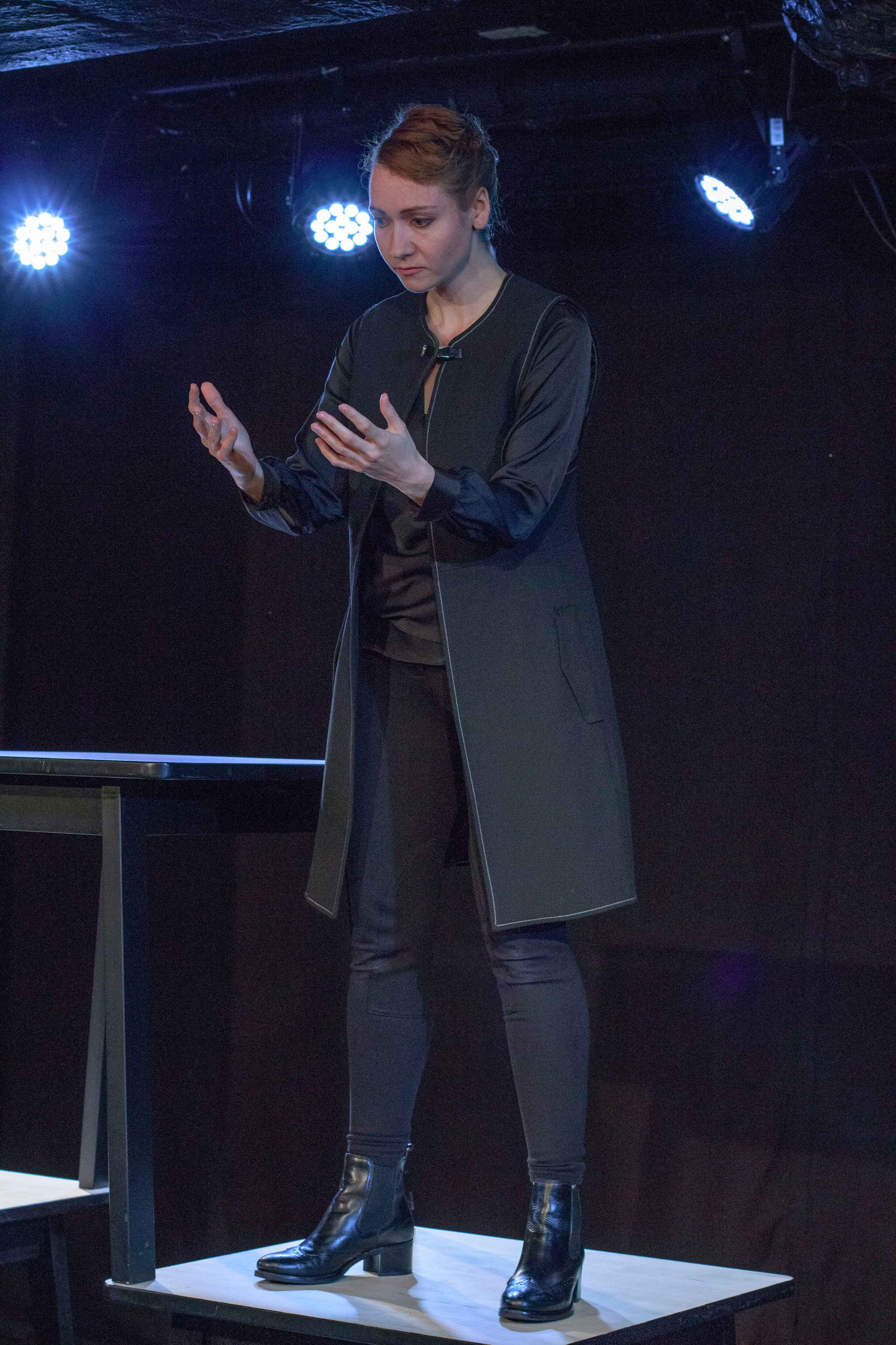 aktorka ubrana na czarno, w czarnym płaszczu stoi na stoliku, trzyma wyciągnięte przed sobą dłonie i patrzy na nie, ciemne tło, pada niebieskie światło