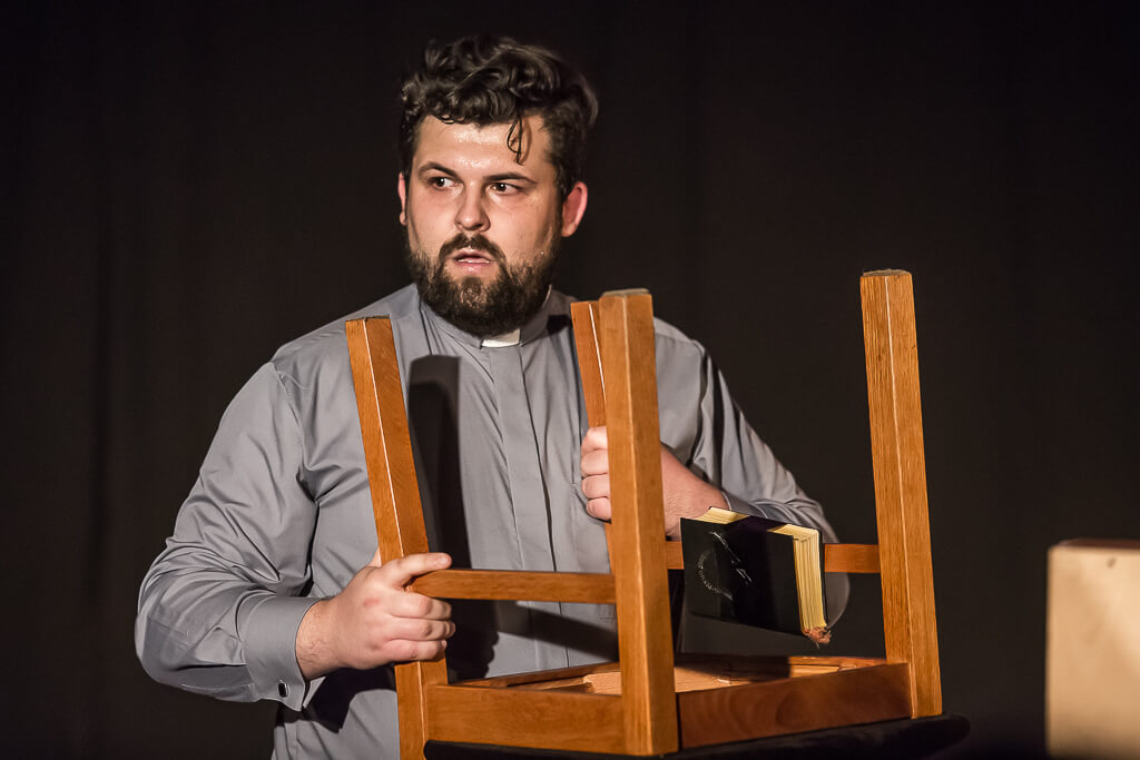 Zdjęcie do spektaklu KAZANIE XXI. Aktor przebrany za księdza trzymający w rękach odwrócone do góry nogami krzesło z leżącą na nim biblią.
