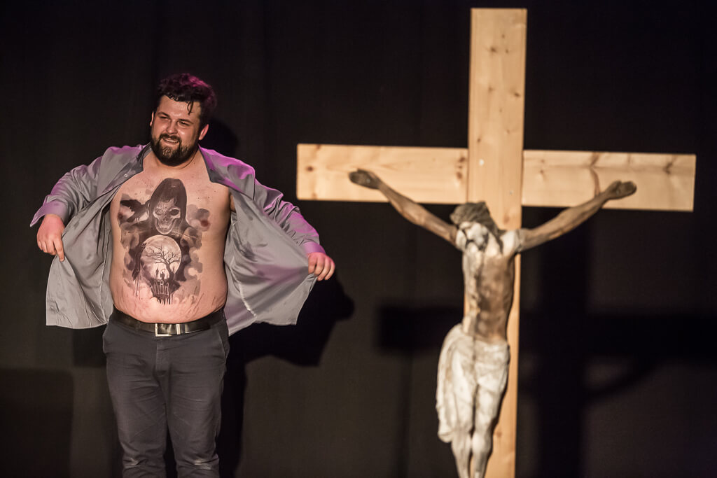 Zdjęcie do spektaklu KAZANIE XXI. Aktor szeroko rozpina swoją koszulę i prezentuje duży tatuaż znajdujący się na jego brzuchu i klatce piersiowej.