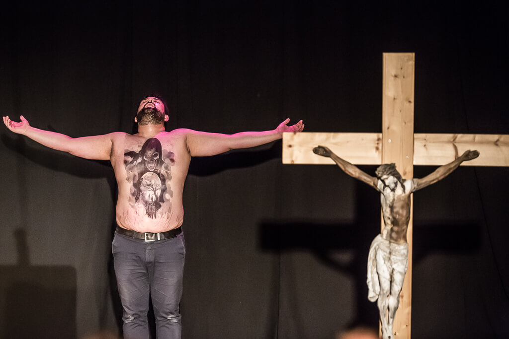 Zdjęcie do spektaklu KAZANIE XXI. Aktor bez koszuli i z dużym tatuażem na swojej klatce piersiowej i brzuchu unosi ręce i zwraca się do Boga. Obok niego stoi duży drewniany krzyż.