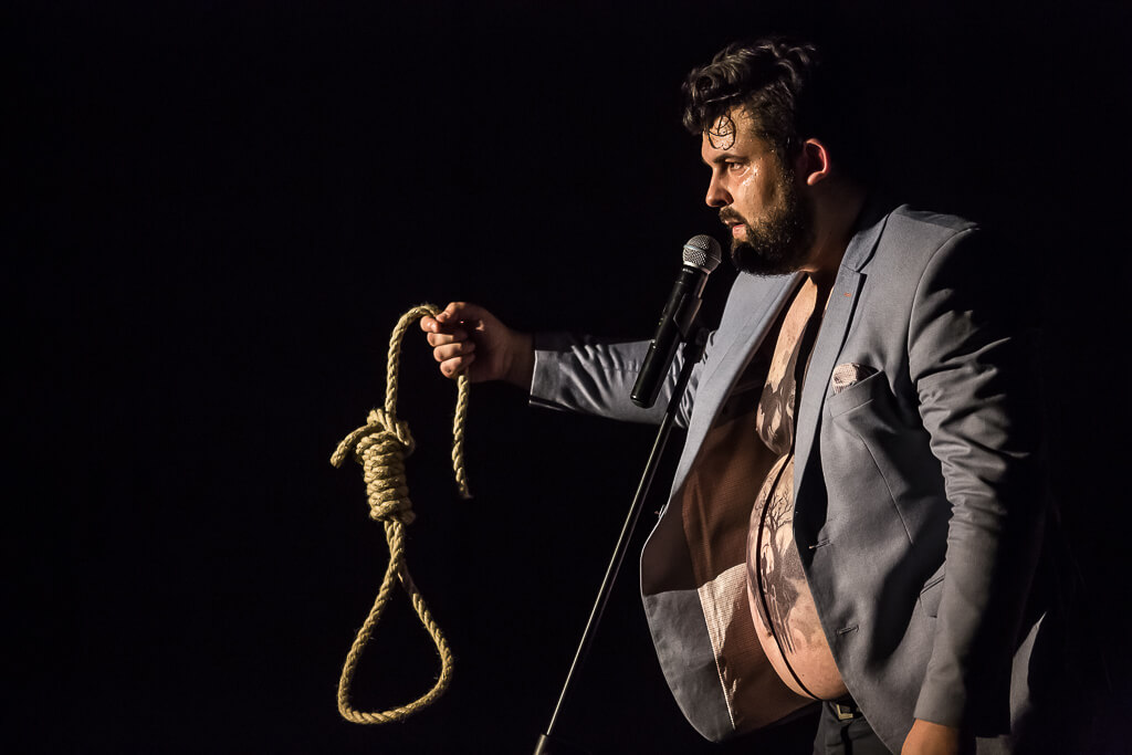 Zdjęcie do spektaklu KAZANIE XXI. Na zdjęciu znajduje się aktor trzymający w ręce pętlę ze sznura, którą prezentuje publice.