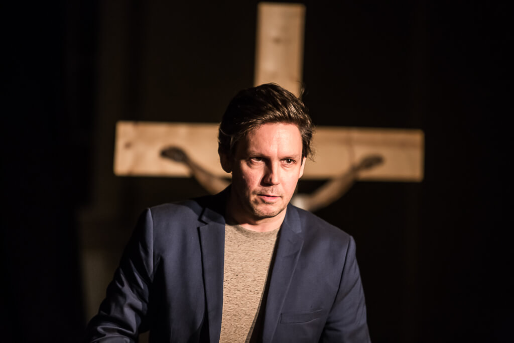 Zdjęcie do spektaklu KAZANIE XXI. Na zdjęciu widać elegancko ubranego aktora z poważnym grymasem na twarzy i stojący za nim duży drewniany krzyż.