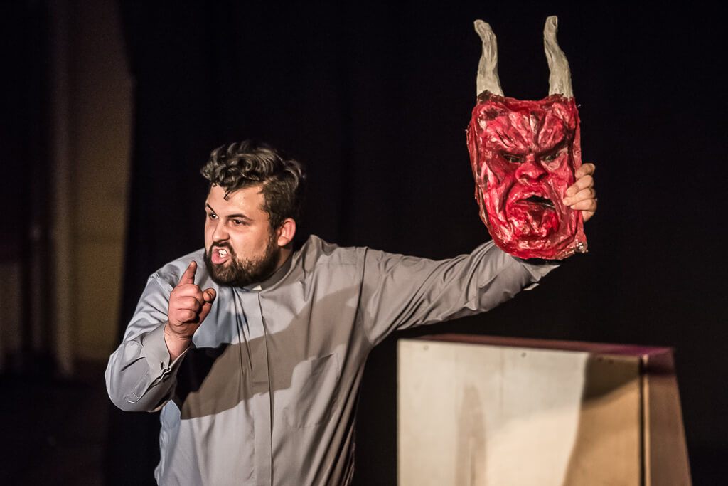 Zdjęcie do spektaklu KAZANIE XXI. Aktor przebrany za księdza trzyma w ręce maskę z podobizną diabła i z zdenerwowaniem na twarzy wypowiada swoją kwestię w stronę publiczności.