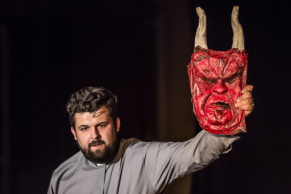 Zdjęcie do spektaklu KAZANIE XXI. Aktor ubrany w koszulę, trzymający w ręce maskę przypominającą diabła.