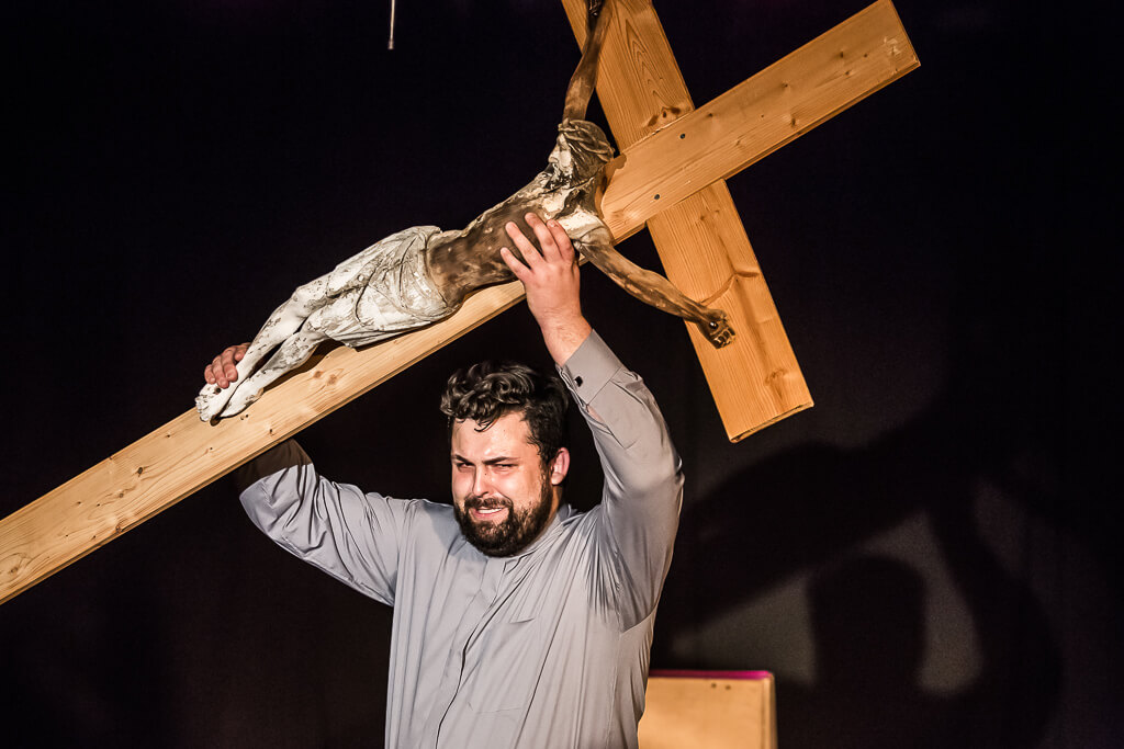 Zdjęcie do spektaklu KAZANIE XXI. Aktor z widocznym wysiłkiem na twarzy trzyma podniesiony w górę krzyż.