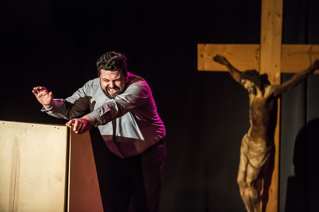 Zdjęcie do spektaklu KAZANIE XXI. Aktor stojący przy mównicy śmieje się, a za nim stoi duży drewniany krzyż z podobizną ukrzyżowanego na nim Jezusa.