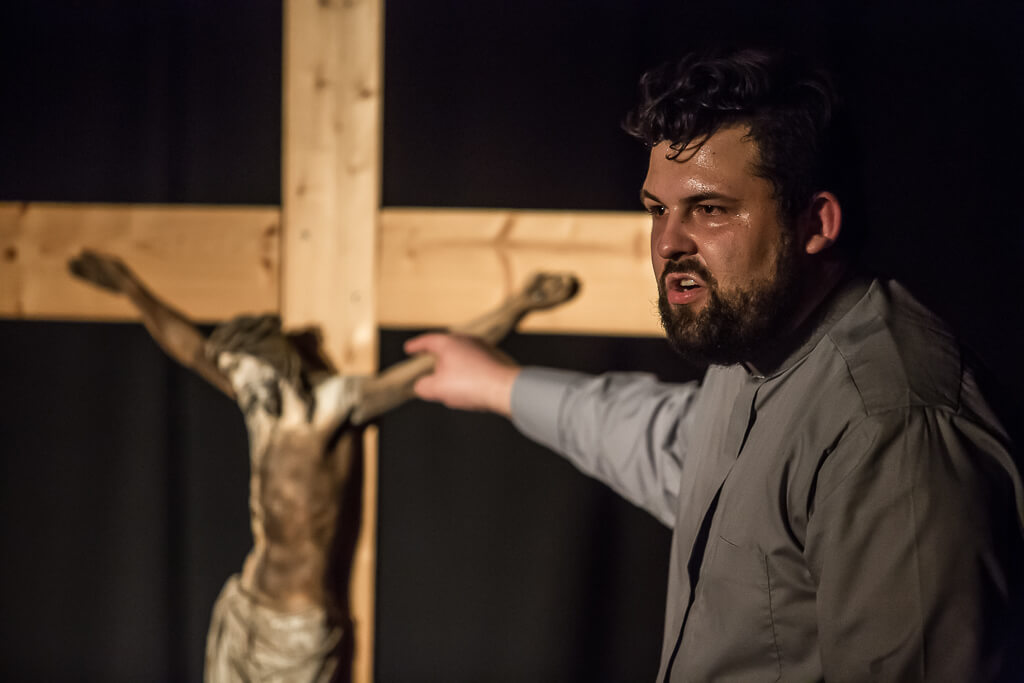 Zdjęcie do spektaklu KAZANIE XXI. Aktor z widocznym na twarzy zmęczeniem wskazuje palcem na stojący za nim duży drewniany krzyż.