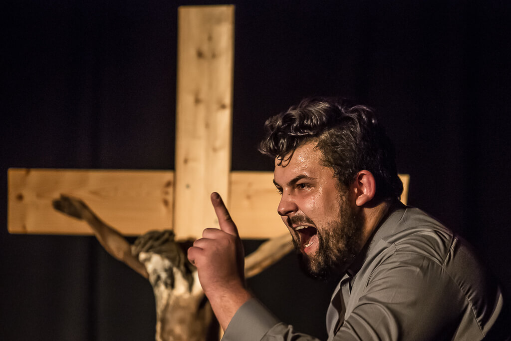 Zdjęcie do spektaklu KAZANIE XXI. Aktor z widocznym zmęczeniem na twarzy i uniesionym palcem wygłasza swoją kwestię, a w tle widać duży drewniany krzyż.