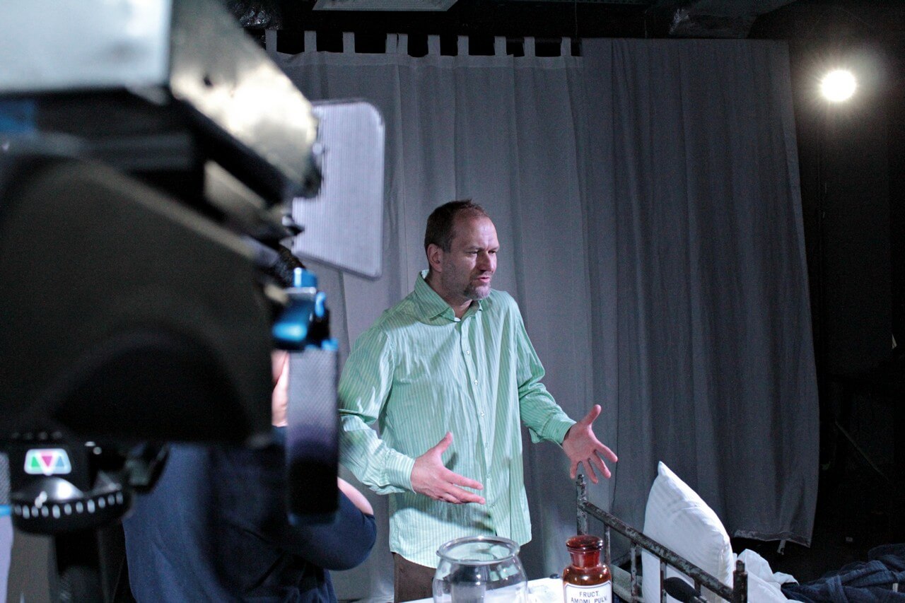 aktor stoi na środku w zielonej koszuli, skierowany jest w prawo, ręce trzyma przed sobą, po lewej stronie widoczna kamera, w tle biała zasłona