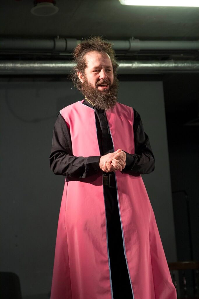 aktor stoi na środku, ubrany na czarno, ma różowy fartuch, dłonie trzy złączone przed sobą