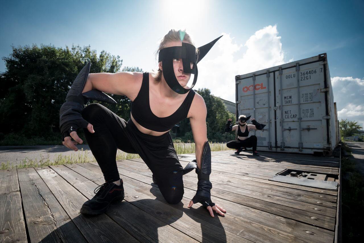 Zdjęcie do spektaklu Masska. Aktor w czarnym kostiumie i masce na twarzy, klęczy na platformie wagonu.