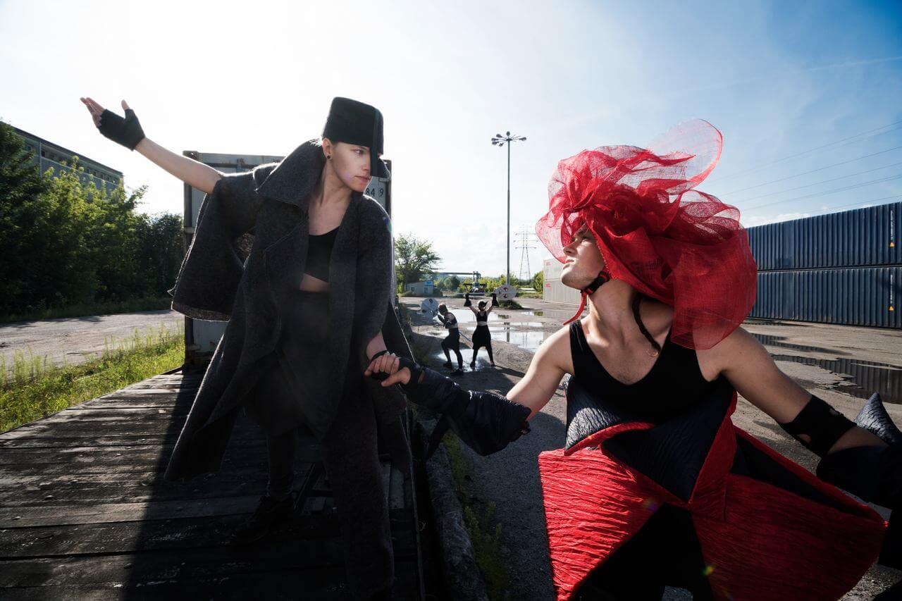 Zdjęcie do spektaklu Masska. Aktorka w czarnym kostiumie i aktor w czarno czerwonym kostiumie trzymają się za rękę. Zdjęcie na platwormie wagonu kolejowego.