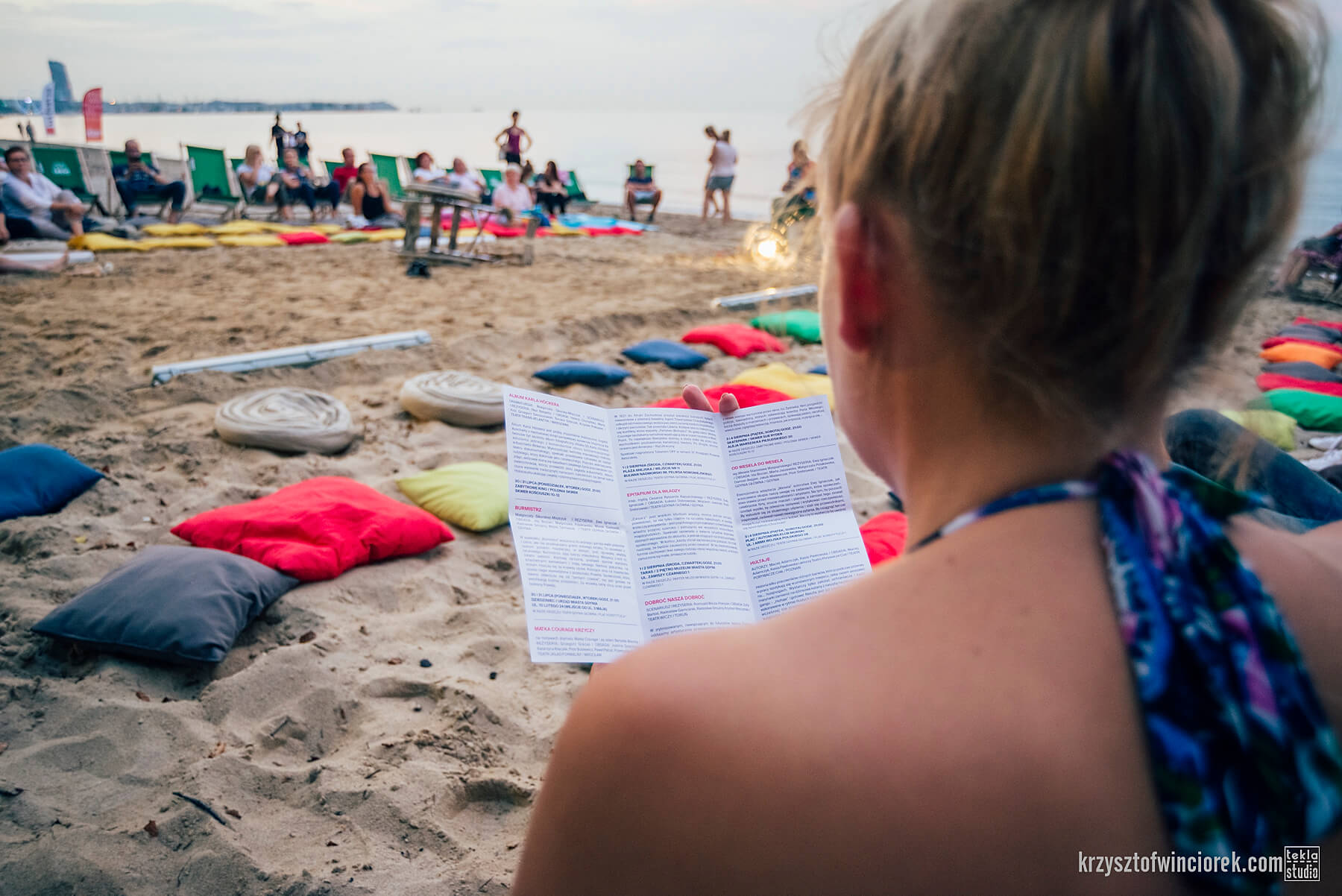 Plaża. Zdjęcie zrobione zza ramienia kobiety, która coś czyta. Na dalszym planie poduchy rozłożone na piasku oraz morze.