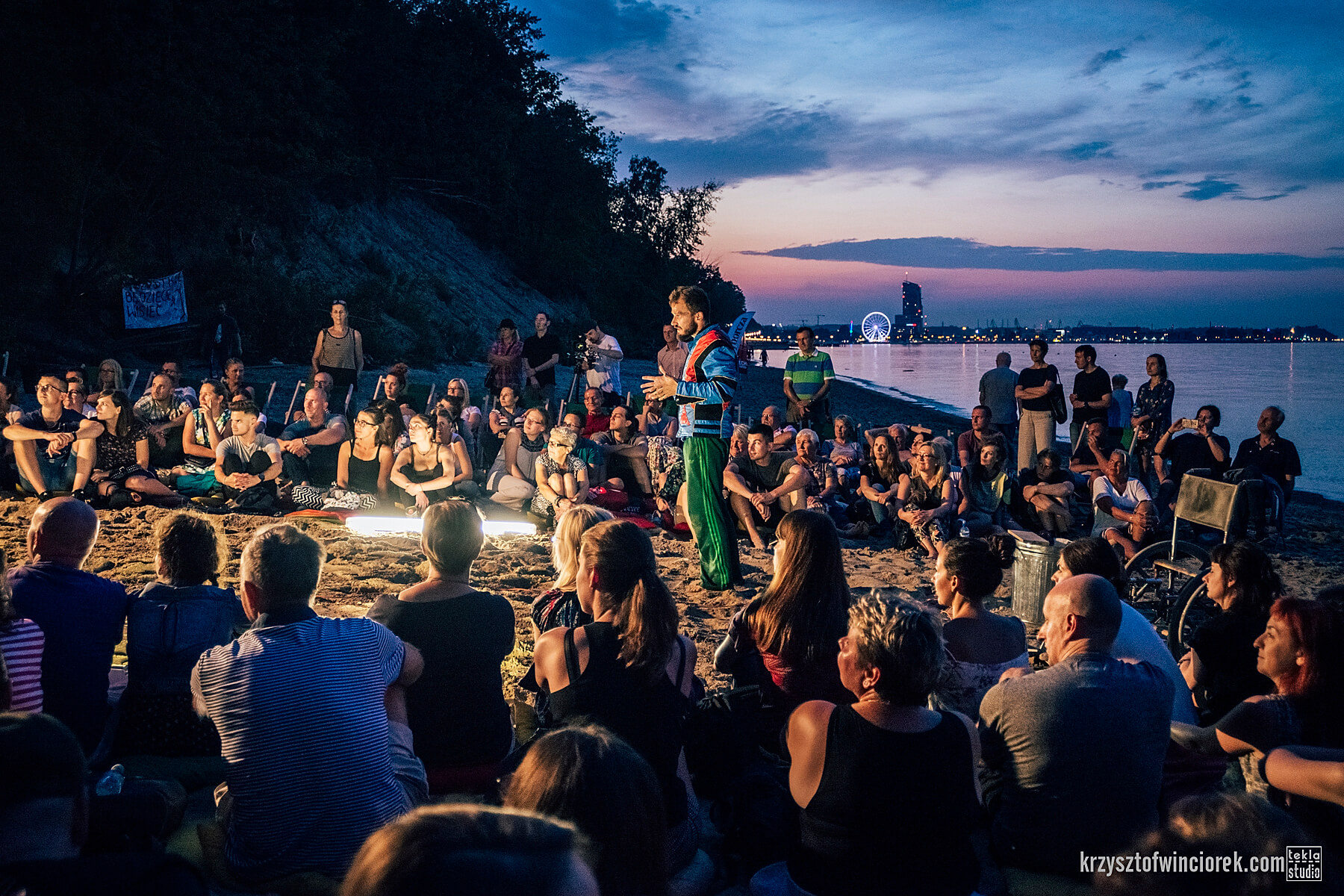 Na zdjęciu duża grupa ludzi siedząca na plaży w okręgu. W jego środku stoi aktor z zarostem ubrany w zielone spodnie i niebiesko-czarny sweter. W oddali widać światła centrum Gdyni.