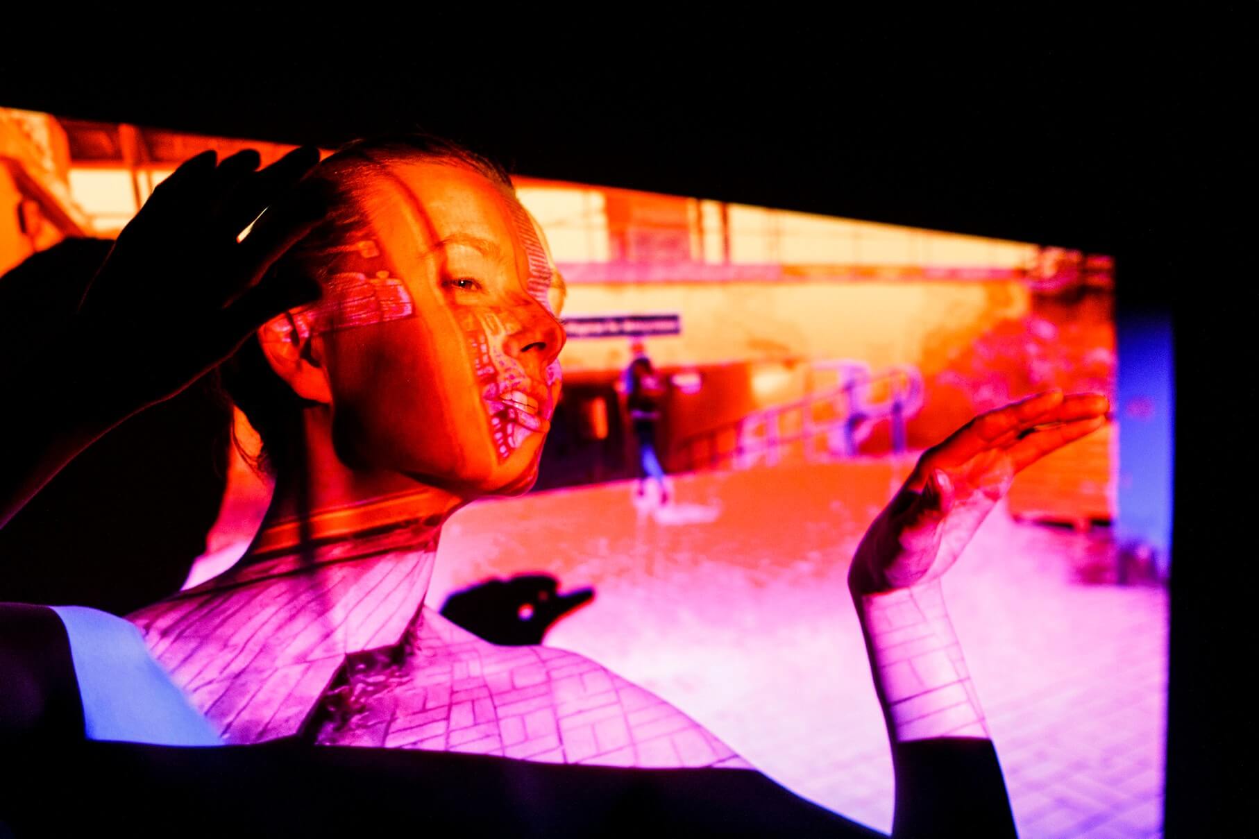 aktorka po lewej stronie oparta o ścianę, zwrócona w prawo, dłonie podniesione na wysokość szyi, ma lekko otwarte usta i zmrużone oczy, pada na nią pomarańczowo fioletowy obraz z projektora