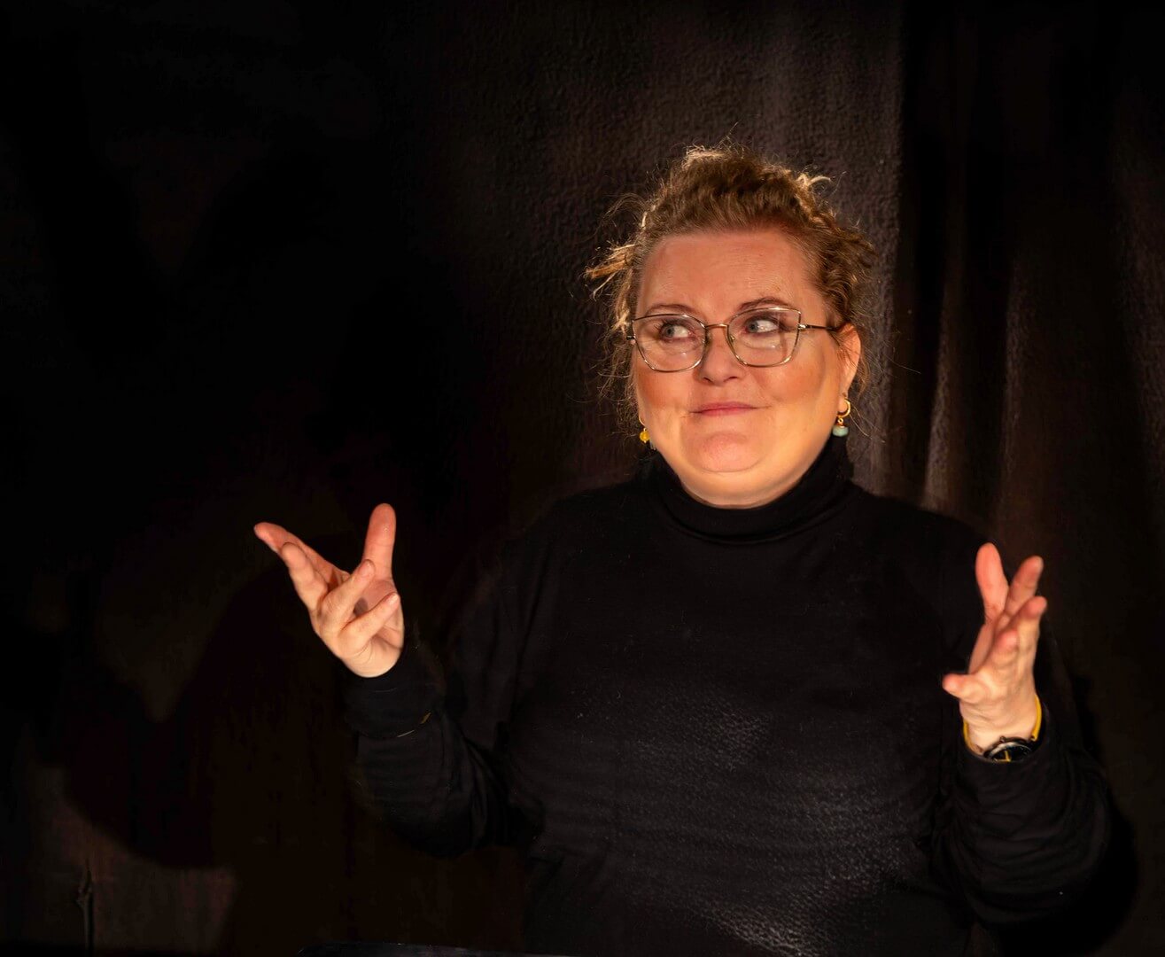 Na zdjęciu kobieta ubrana na czarno, na czarnym tle z uniesionymi przed siebie dłońmi z rozpostartymi palacami