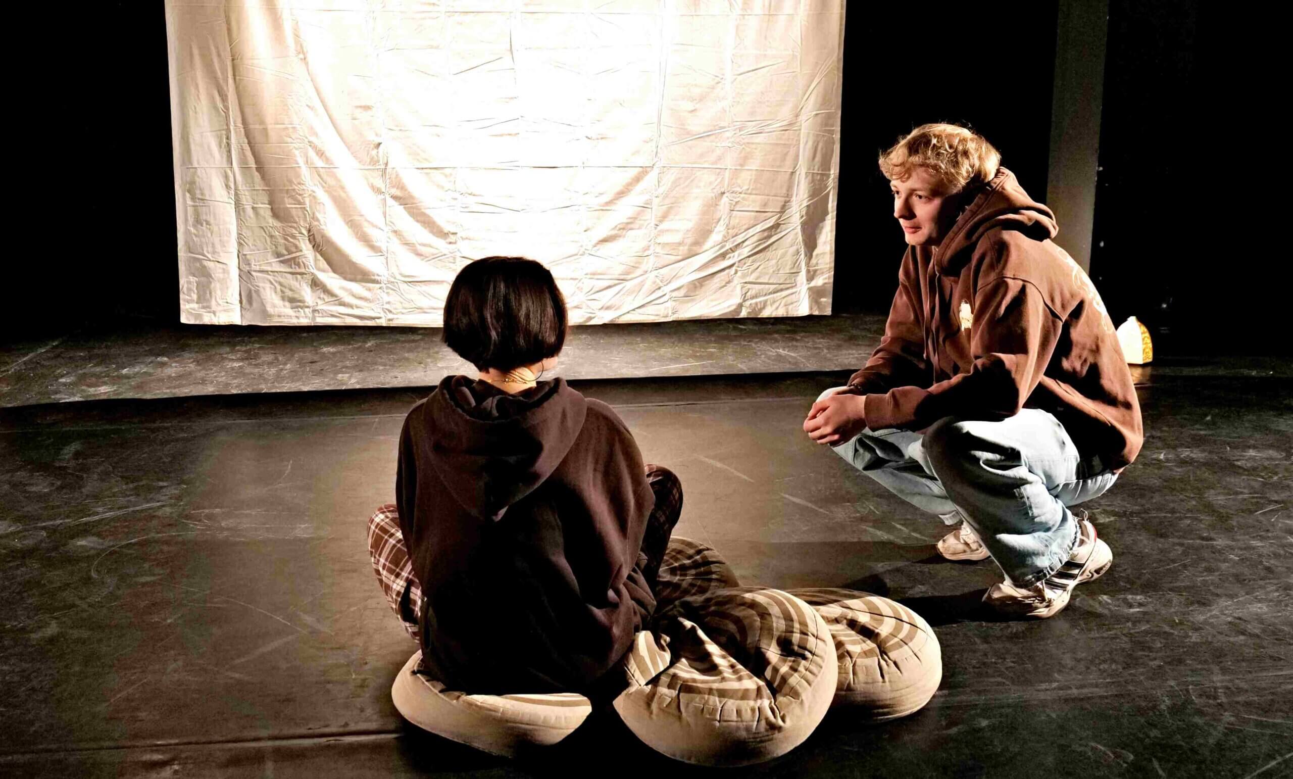 Na zdjęciu dwoje aktorów, aktorka siedzi tyłem na okrągłych poduszkach, obok z prawej kuca aktor. w tle wisi biały ekran