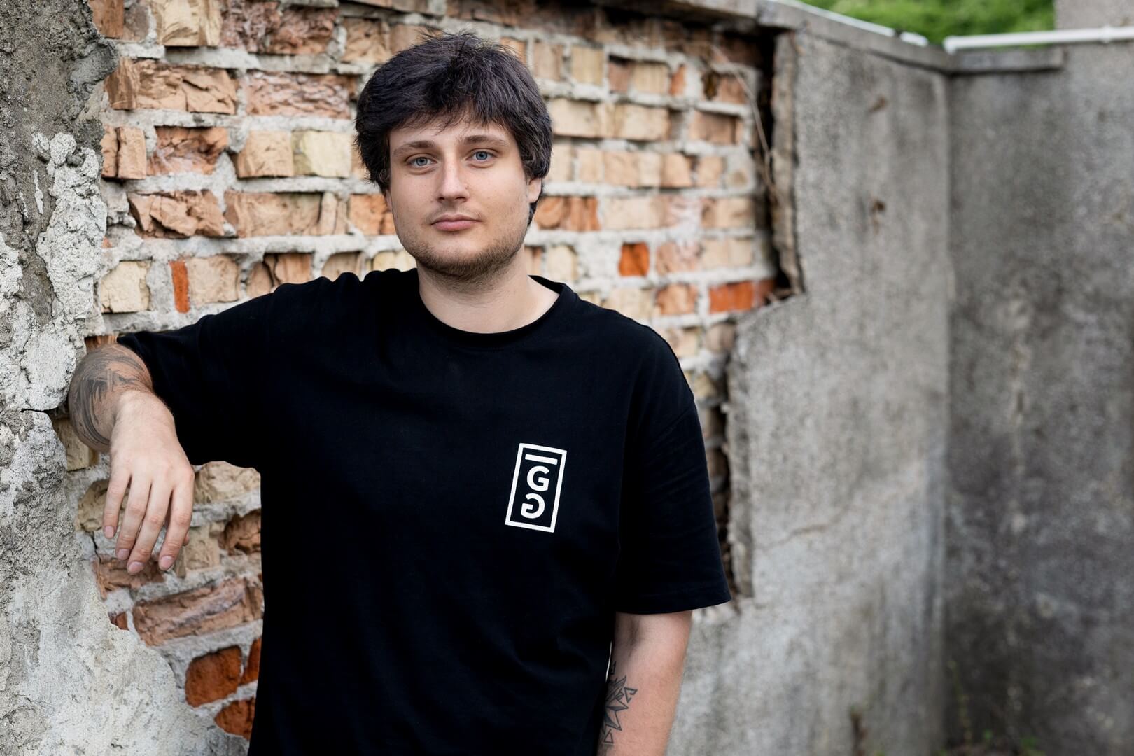 Młody mężczyzna o ciemnych włosach, w czarnym T-shircie z białym logo, opiera się o zniszczoną ścianę z widocznymi cegłami.