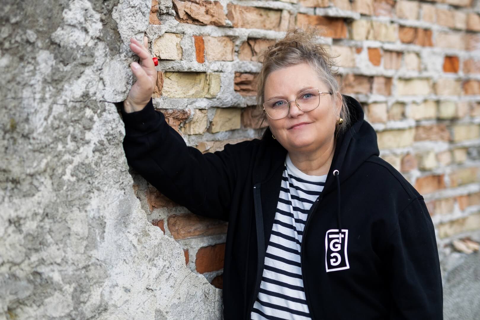 Na zdjęciu kobieta w okularach, czarnej bluzie z kapturem i koszulce w czarno-białe paski stoi przy starym ceglanym murze. Jedną ręką opiera się o mur i patrzy w stronę aparatu z lekkim uśmiechem