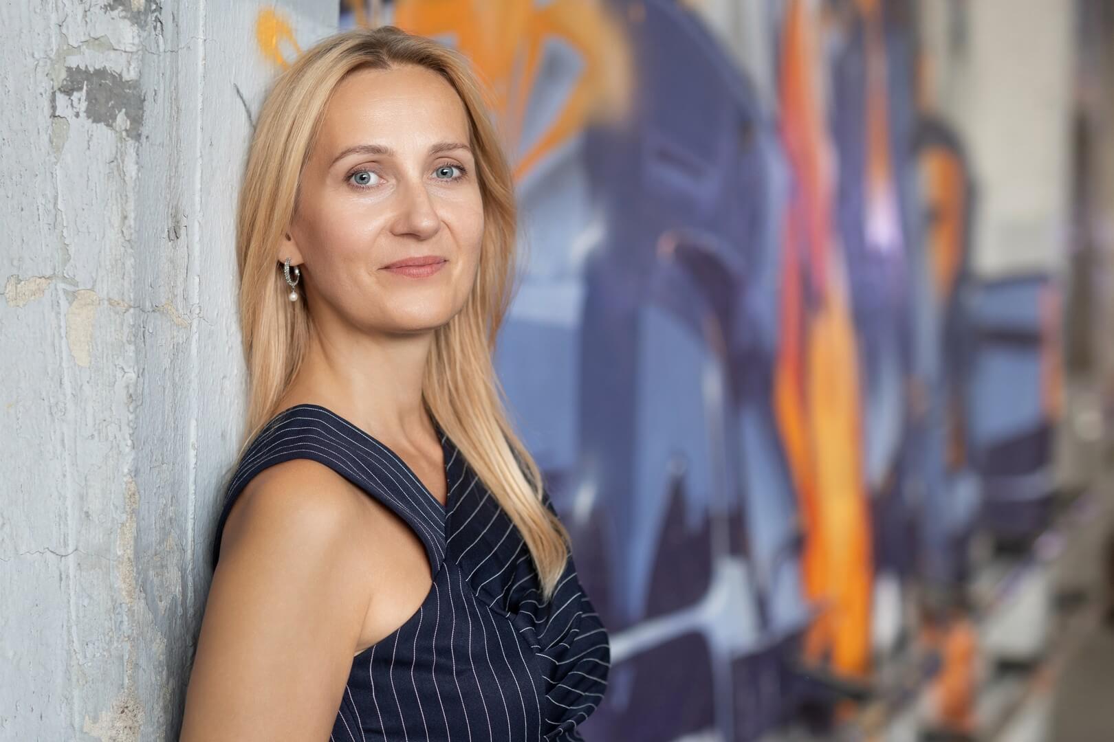 Portret uśmiechniętej, blondynki w ciemnej sukience w prążki, opierającej się o betonową ścianę z kolorowym graffiti w tle.