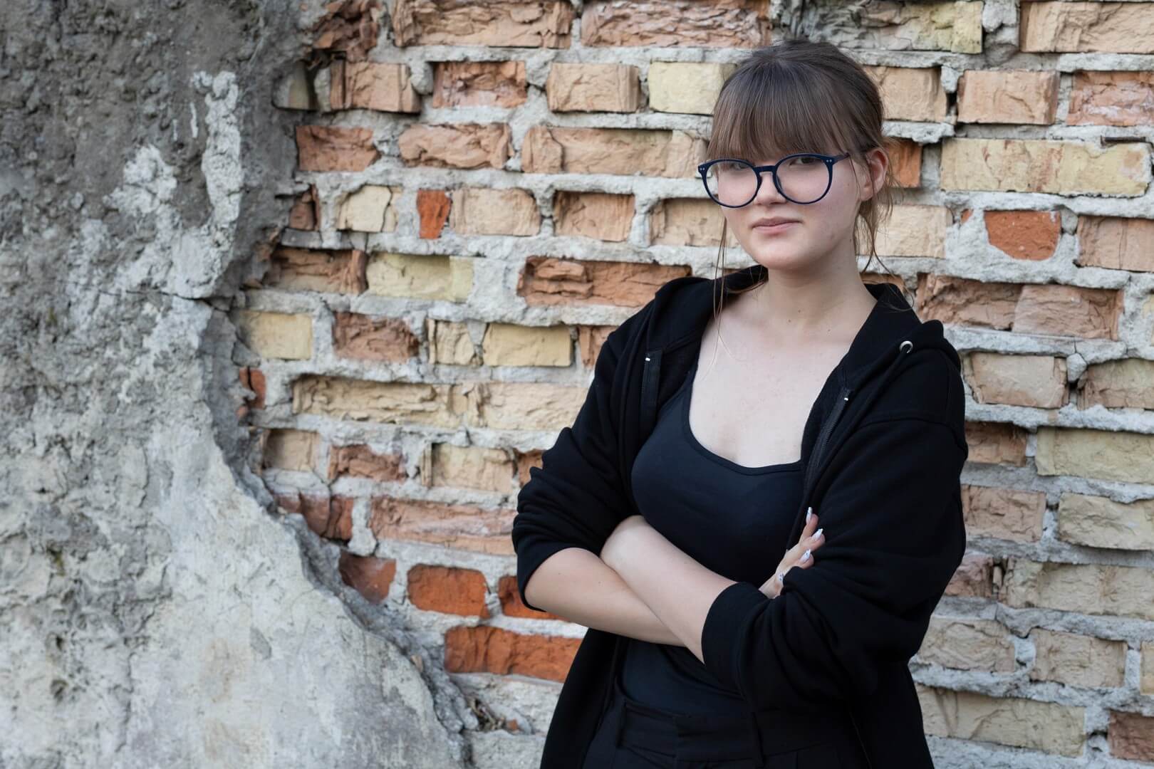 Młoda kobieta w okularach i czarnej rozpinanej bluzie, z rękami skrzyżowanymi na piersi, pozuje na tle zniszczonego muru z cegieł i betonu.
