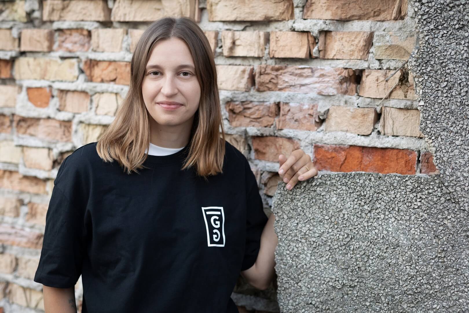 Młoda kobieta o brązowych włosach, w czarnym T-shircie z białym logo, pozuje na tle starej ceglanej ściany.
