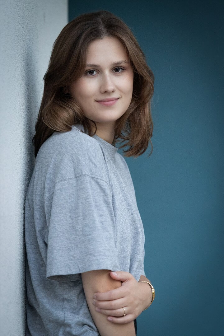Portret młodej kobiety z brązowymi włosami, w szarym T-shircie, opierającej się o jasną ścianę na niebieskim tle.