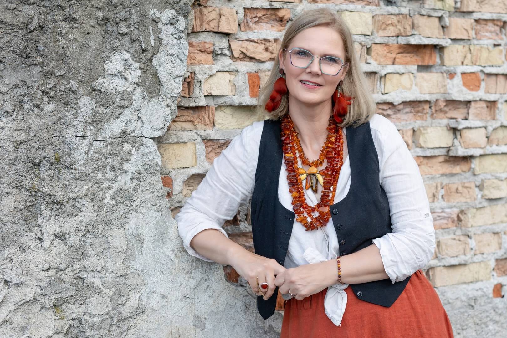 Uśmiechnięta blondynka w okularach, ujęta od pasa w górę, w białej koszuli, ciemnej kamizelce i bogatej bursztynowej biżuterii, opiera się o zniszczony mur z cegieł.