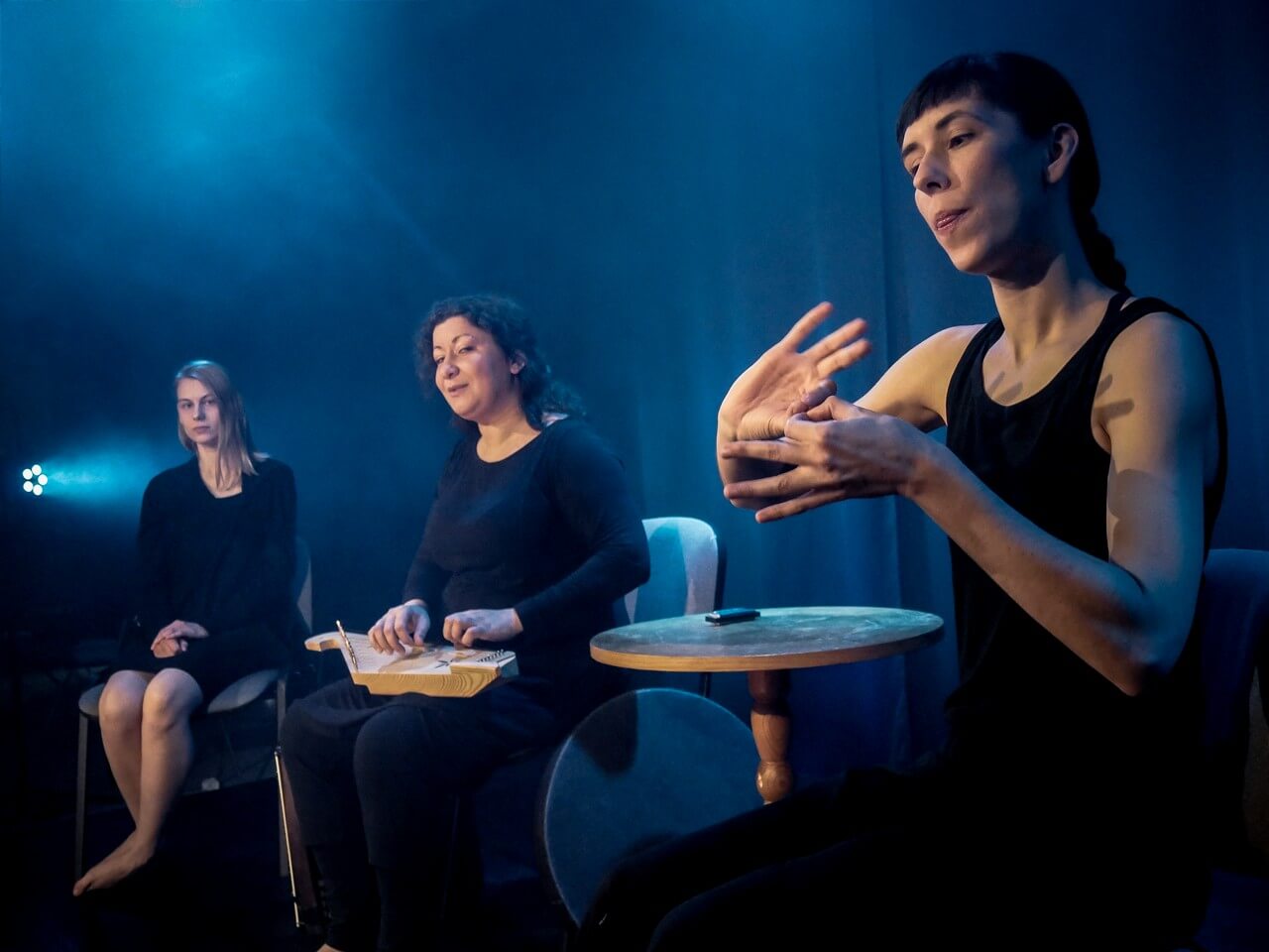 trzy siedzące aktorki ubrane na czarno, kobieta po prawej stronie wykonuje gesty dłońmi, oświetla je niebieskie światło