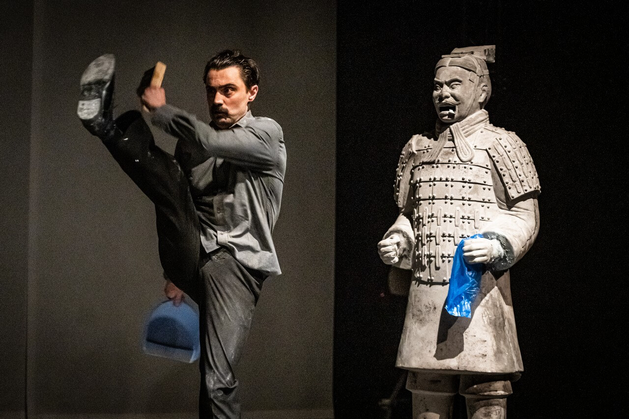 po lewej stronie aktor podnosi nogę do góry, w jednej ręce trzyma szufelkę w drugiej podniesionej szczotkę, po prawej stronie posąg azjatyckiego żołnierza, w lewą dłoń wsadzony niebieski worek foliowy