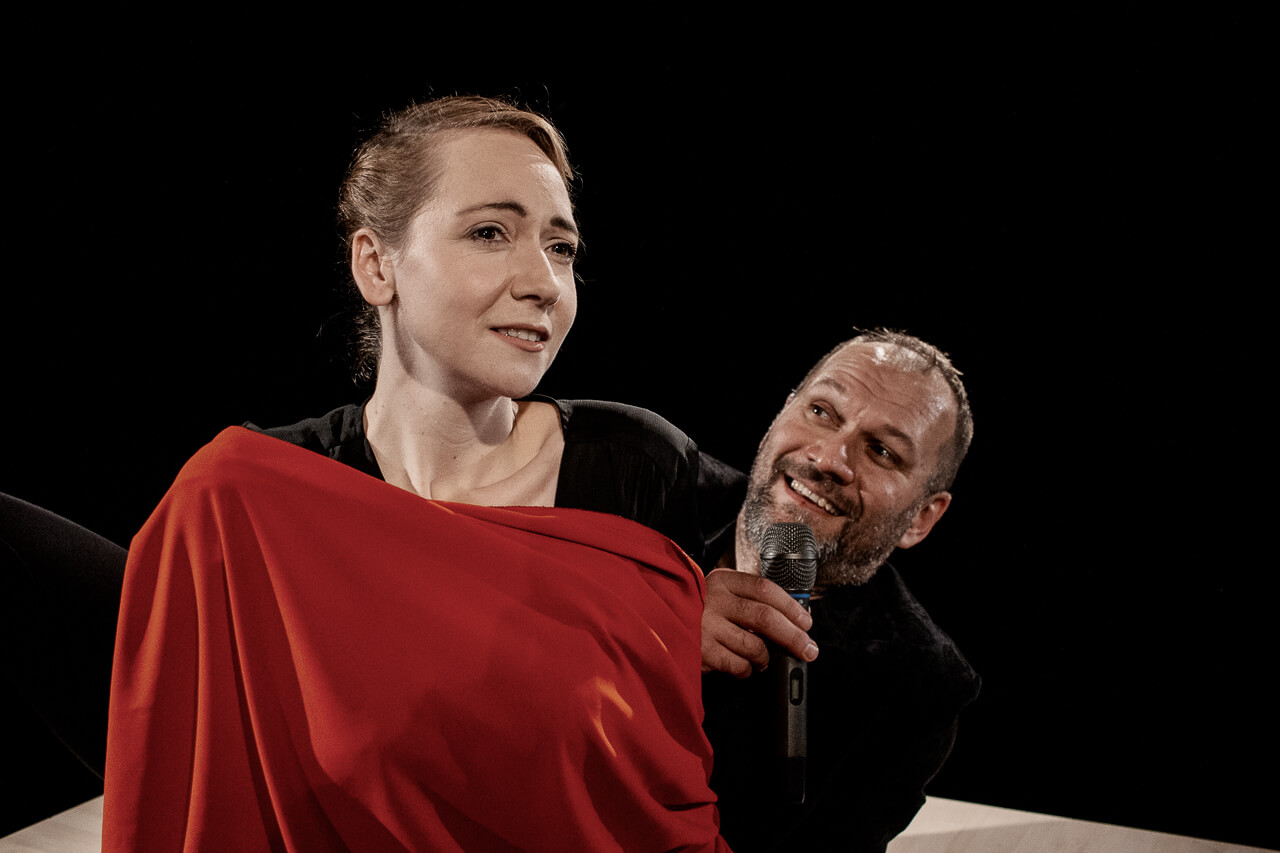 dwoje aktorów, kobieta po lewej stronie zwrócona w lewo okryta czerwonym materiałem, po prawej mężczyzna ubrany na czarno, w prawej ręce trzyma mikrofon blisko ust, czarne tło