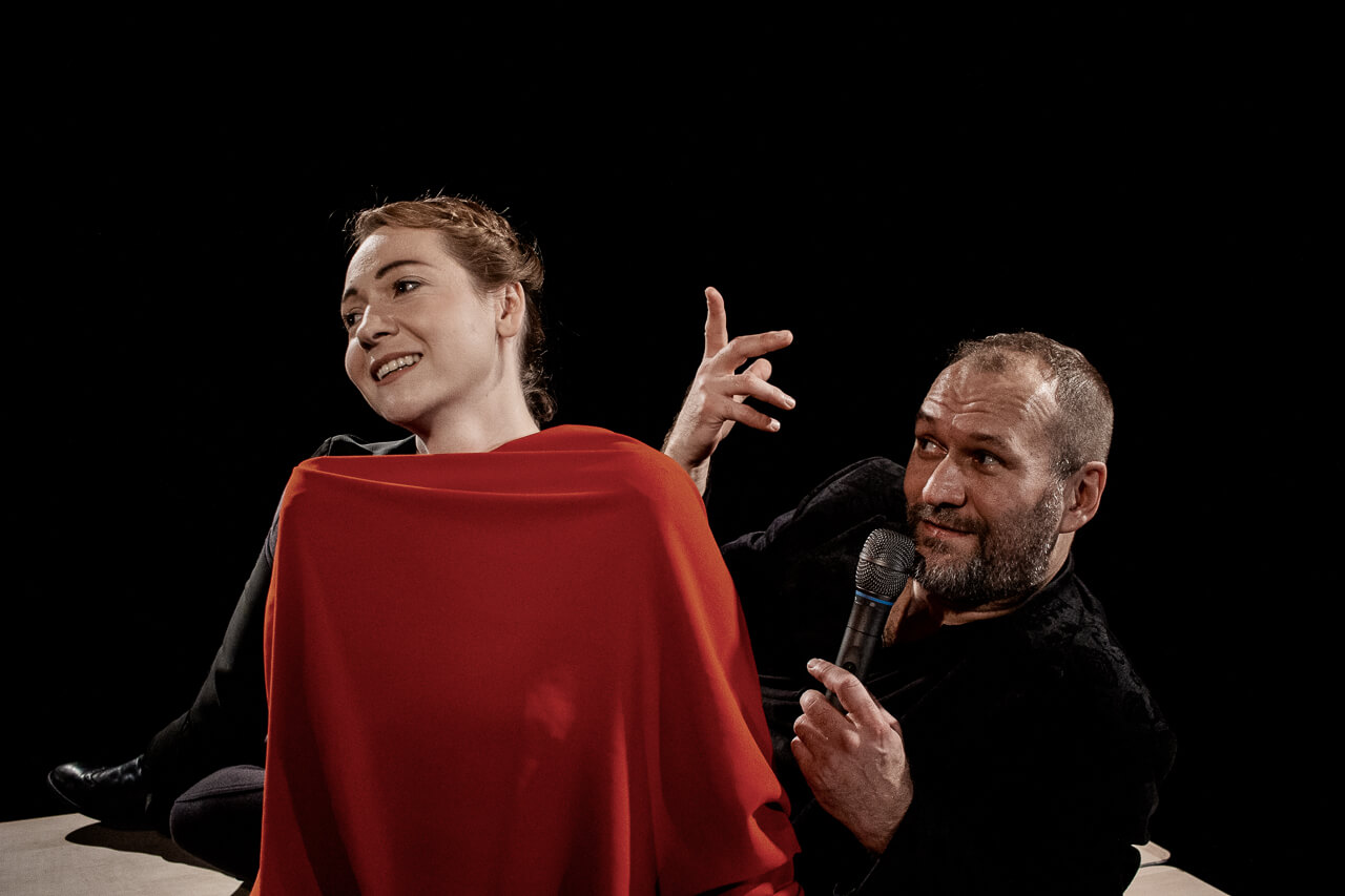 dwoje aktorów, kobieta po lewej stronie zwrócona w lewo okryta czerwonym materiałem, po prawej mężczyzna ubrany na czarno, prawą rękę unosi do góry, w lewe ręce trzyma mikrofon blisko ust, patrzy na kobietę, czarne tło