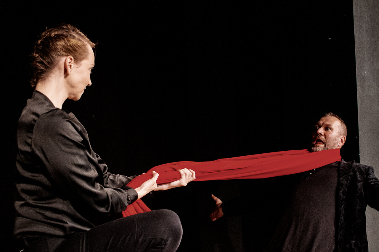 po lewej stronie aktorka ubrana na czarno kuca, zwrócona w prawo, trzyma dłońmi czerwony materiał owinięty wokół szyi drugiego aktora stojącego po prawej, mężczyzna ubrany na czarno odchyla się do tyłu, ma ręce rozłożone na boki i zdziwiony wyraz twarzy