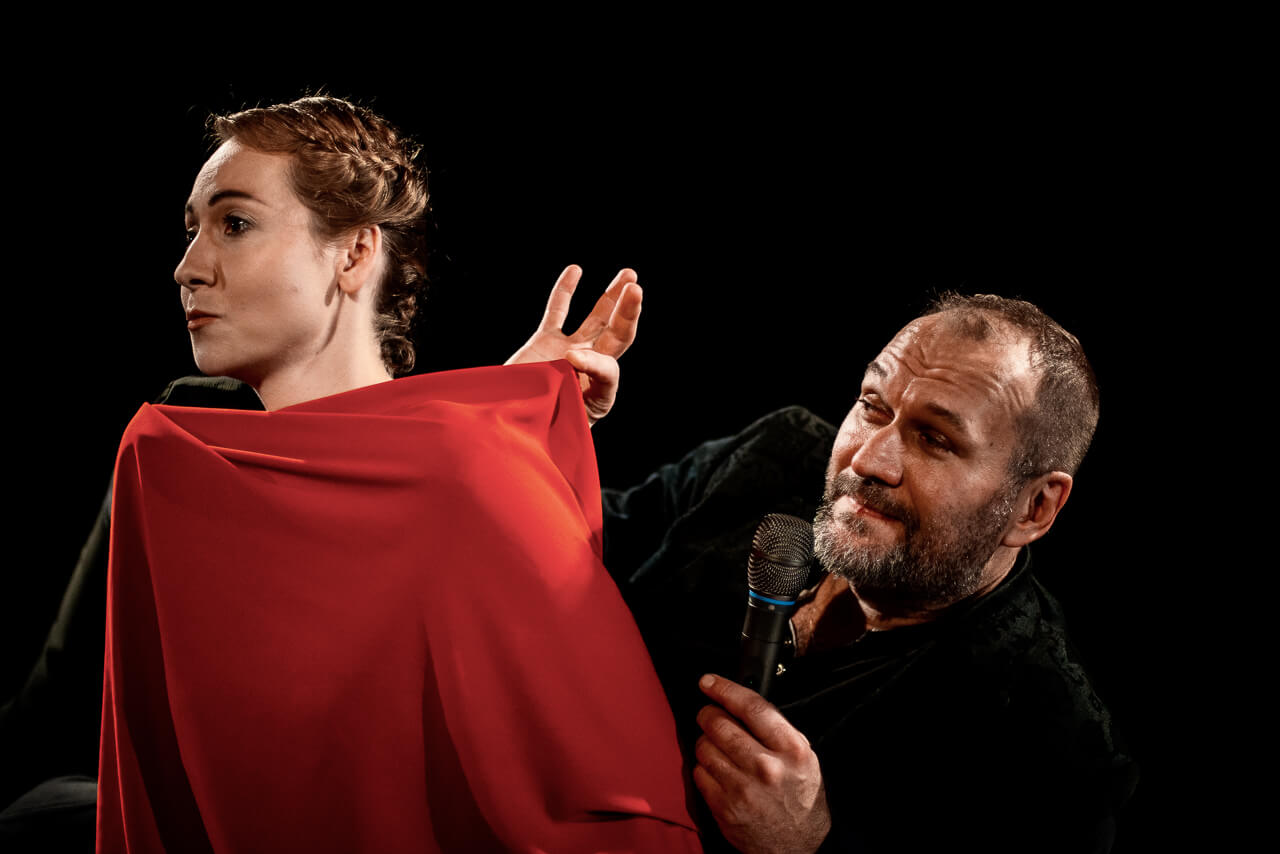 dwoje aktorów, kobieta po lewej stronie zwrócona w lewo okryta czerwonym materiałem, po prawej mężczyzna ubrany na czarno, prawą ręką dotyka czerwonego materiału, w lewe ręce trzyma mikrofon blisko ust, czarne tło