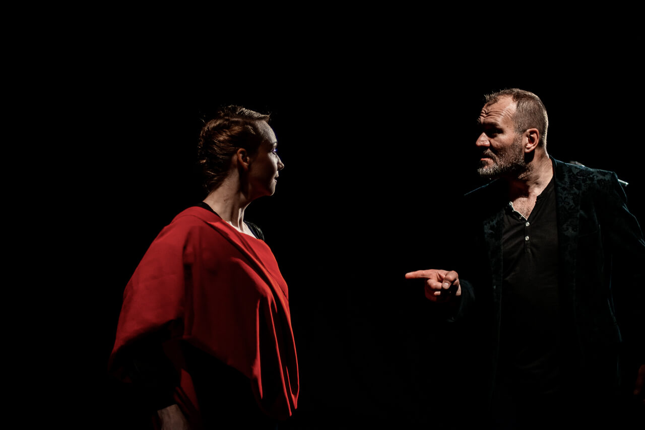 dwoje aktorów, kobieta po lewej stronie zwrócona w prawo, okryta czerwonym materiałem, po prawej mężczyzna ubrany na czarno zwrócony w lewo patrzy na kobietę i wskazuje na nią palcem prawej dłoni, czarne tło