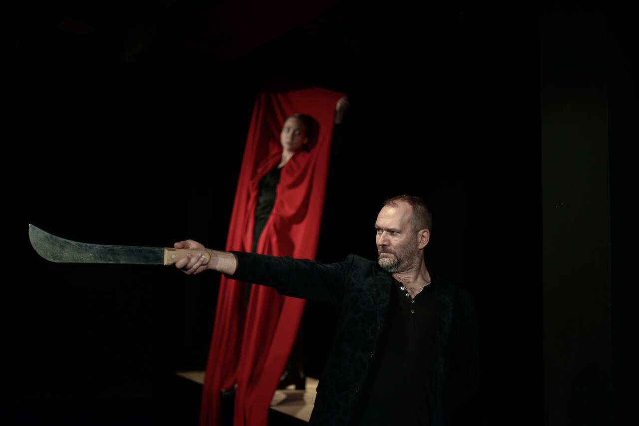dwoje aktorów, na pierwszym planie mężczyzna ubrany na czarno zwrócony w lewo, prawą rękę ma wyciągniętą do przodu, trzyma w niej ostre narzędzie, w tle stoi kobieta z rękami wyciągniętymi do góry w których trzyma obszerny czerwony materiał okalający jej postać, czarne tło