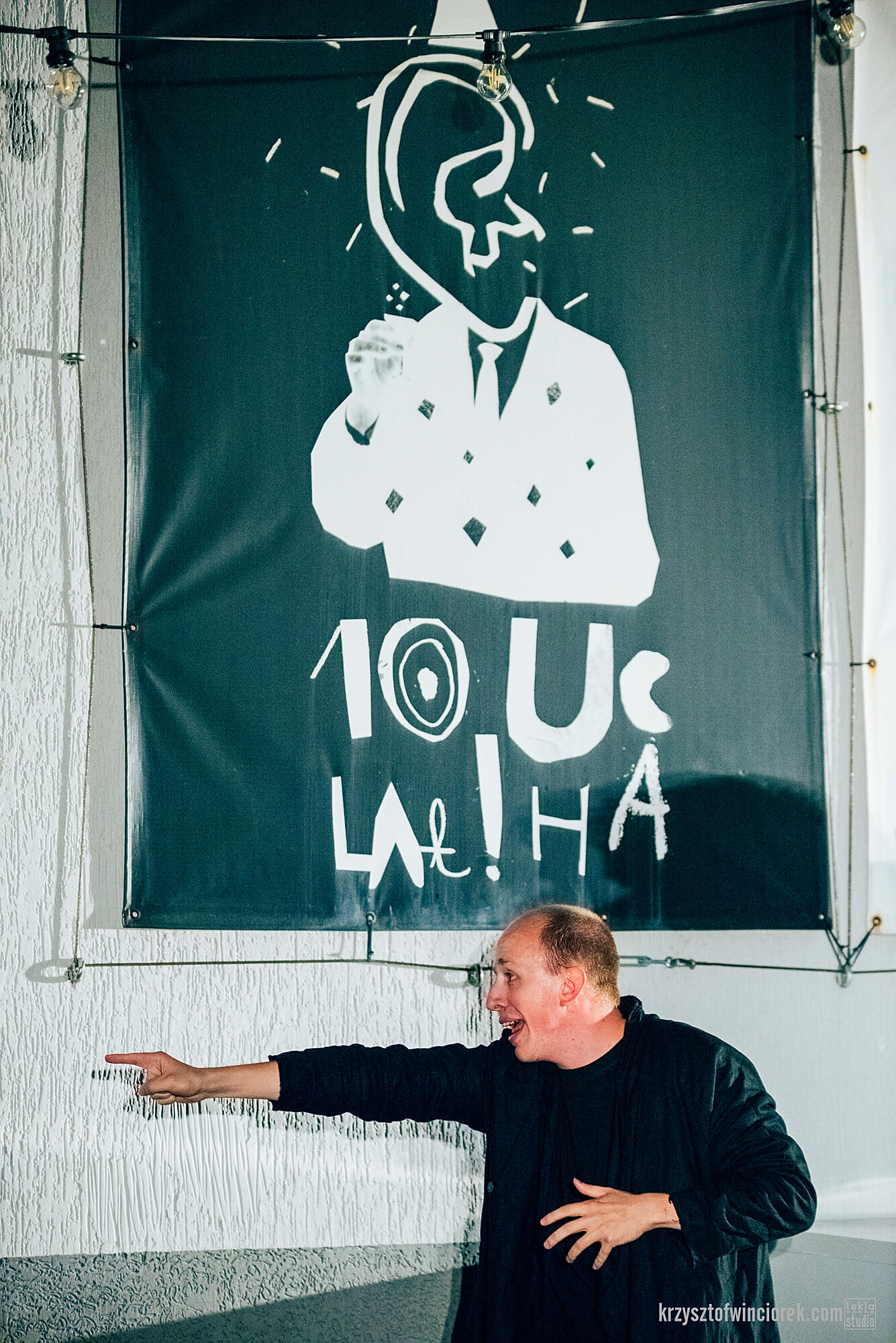 Zdjęcie z festiwalu Pociąg do miasta. Aktor z wyciągniętą w lewo reką i wskazującym palcem. Za nim na ścianie baner z napisem 10 lat ucha.