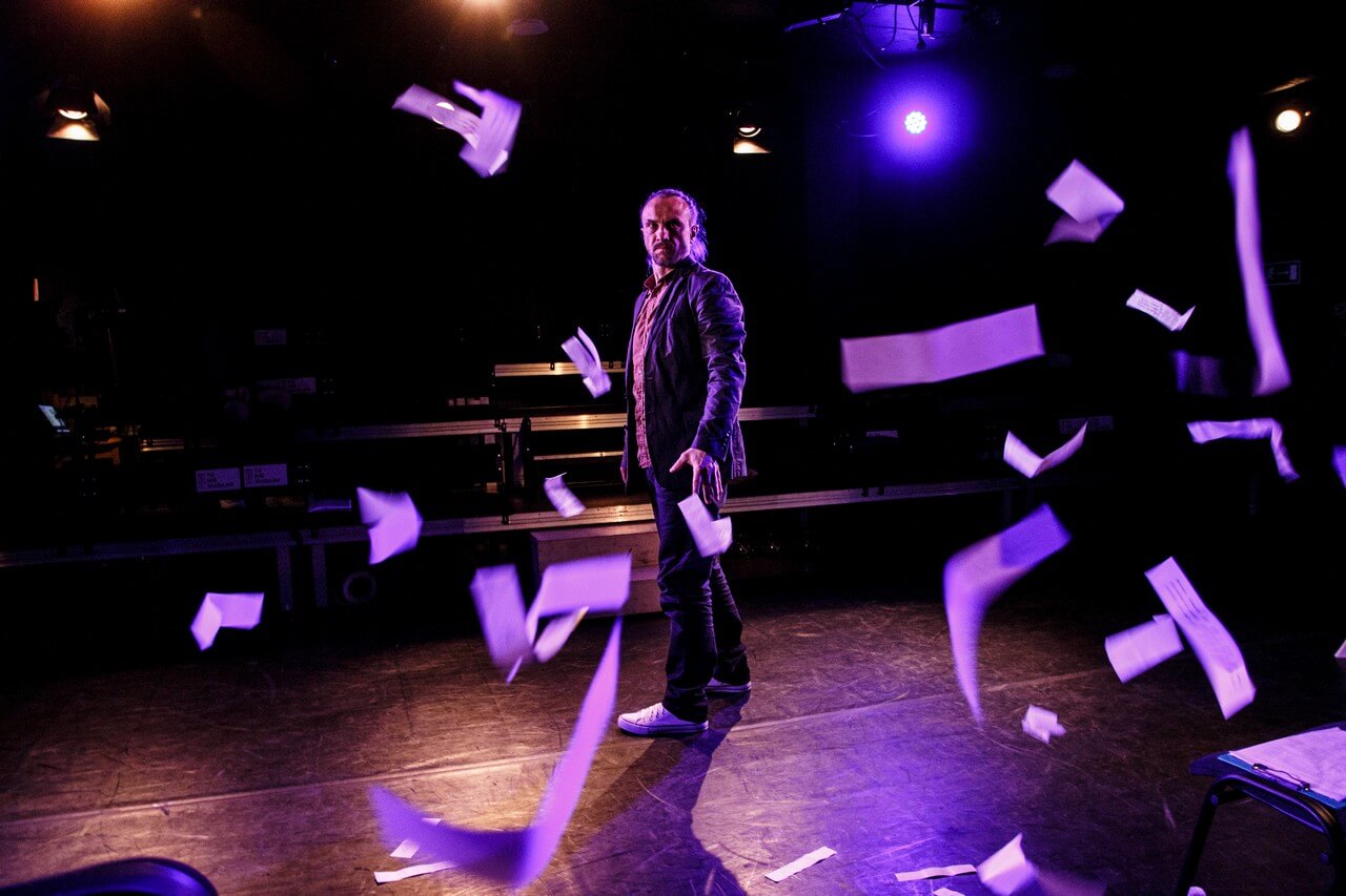 aktor stoi na scenie na środku, dookoła niego spadają rozrzucone fragmenty kartek, pada fioletowe światło