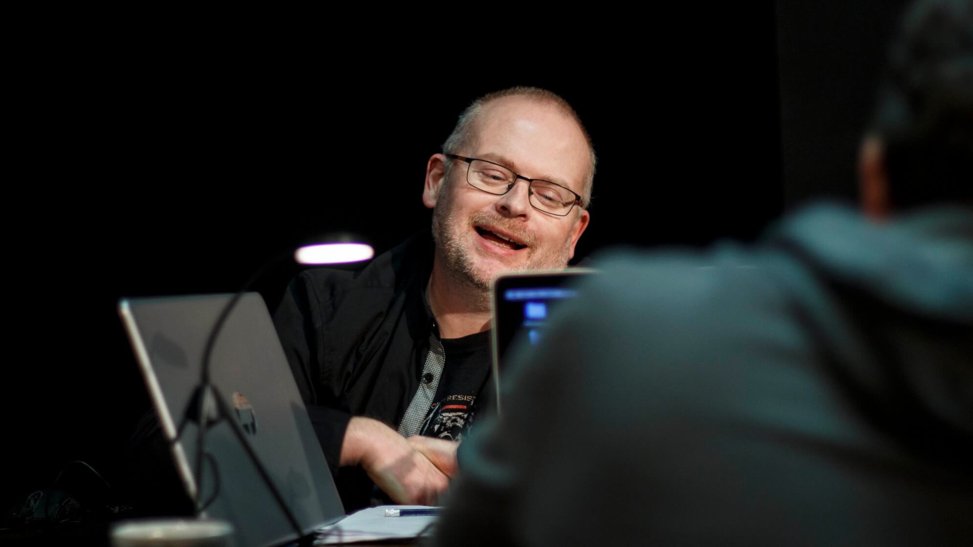 mężczyzna na środku w okularach ubrany na czarno, uśmiecha się, siedzi przy stoliku na którym stoi laptop