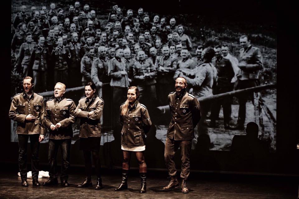 Trzech mężczyzn i dwie kobiety ubrani w mundury SS. Stoją w szeregu Śpiewają. Za nimi wyświetlana projekcja zgromadzonych mężczyzn.