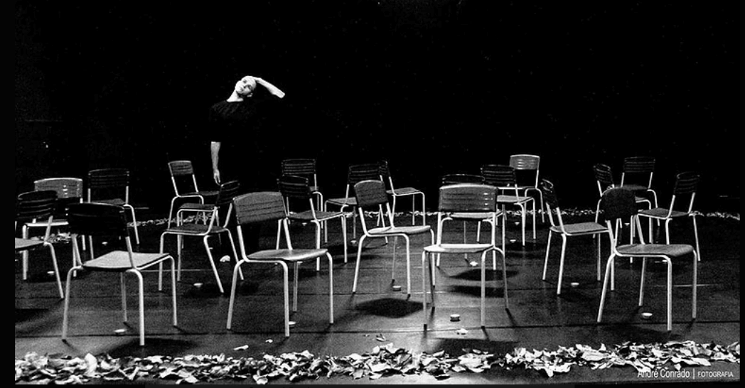 Czarno-białe zdjęcie z Festiwalu Between.Pomiędzy 2018. Po całej scenie zostały chaotycznie rozmieszczone krzesła, pod którymi stoją lampiony. W tle aktor ubrany na czarno trzymający się lewą ręką za głowę.