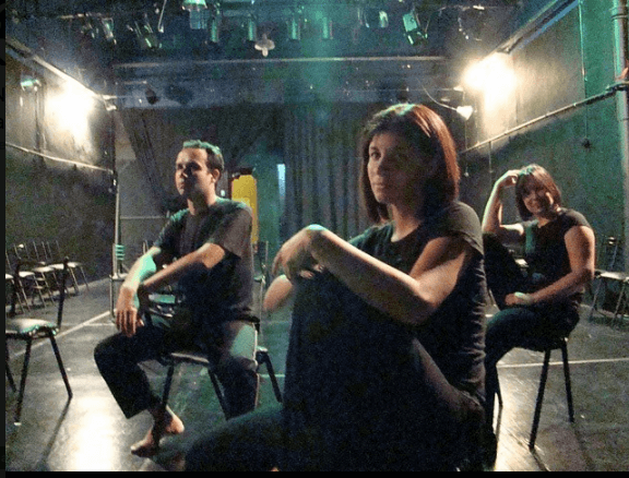 Zdjęcie z Festiwalu Between.Pomiędzy 2018. Na scenie siedzi trójka aktorów. Dwie kobiety i mężczyzna. Kobieta na froncie patrzy w obiektyw aparatu.