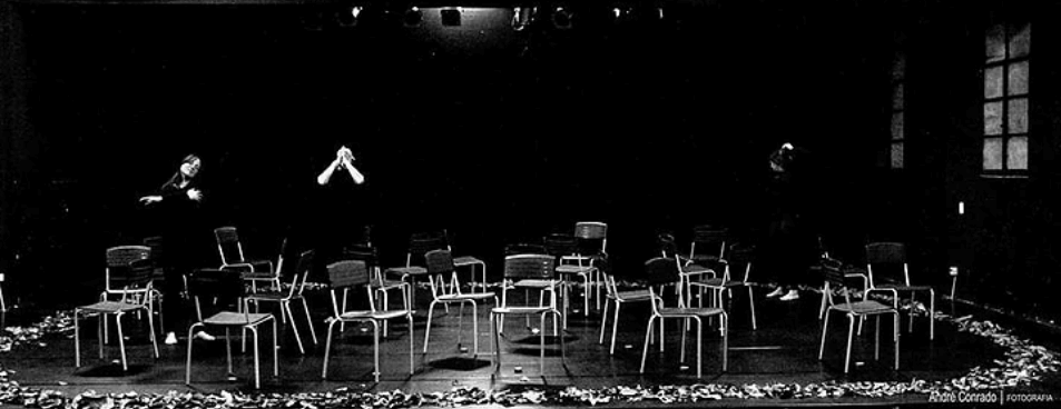 Czarno-białe zdjęcie z Festiwalu Between.Pomiędzy 2018. Chaotycznie ustawione na scenie krzesła, pod którymi stoją lampiony. W tle widać dwójkę aktorów, kobietę i mężczyznę, ubranych na czarno. Kobieta wykonuje taniec, a mężczyzna trzyma się za głowę.