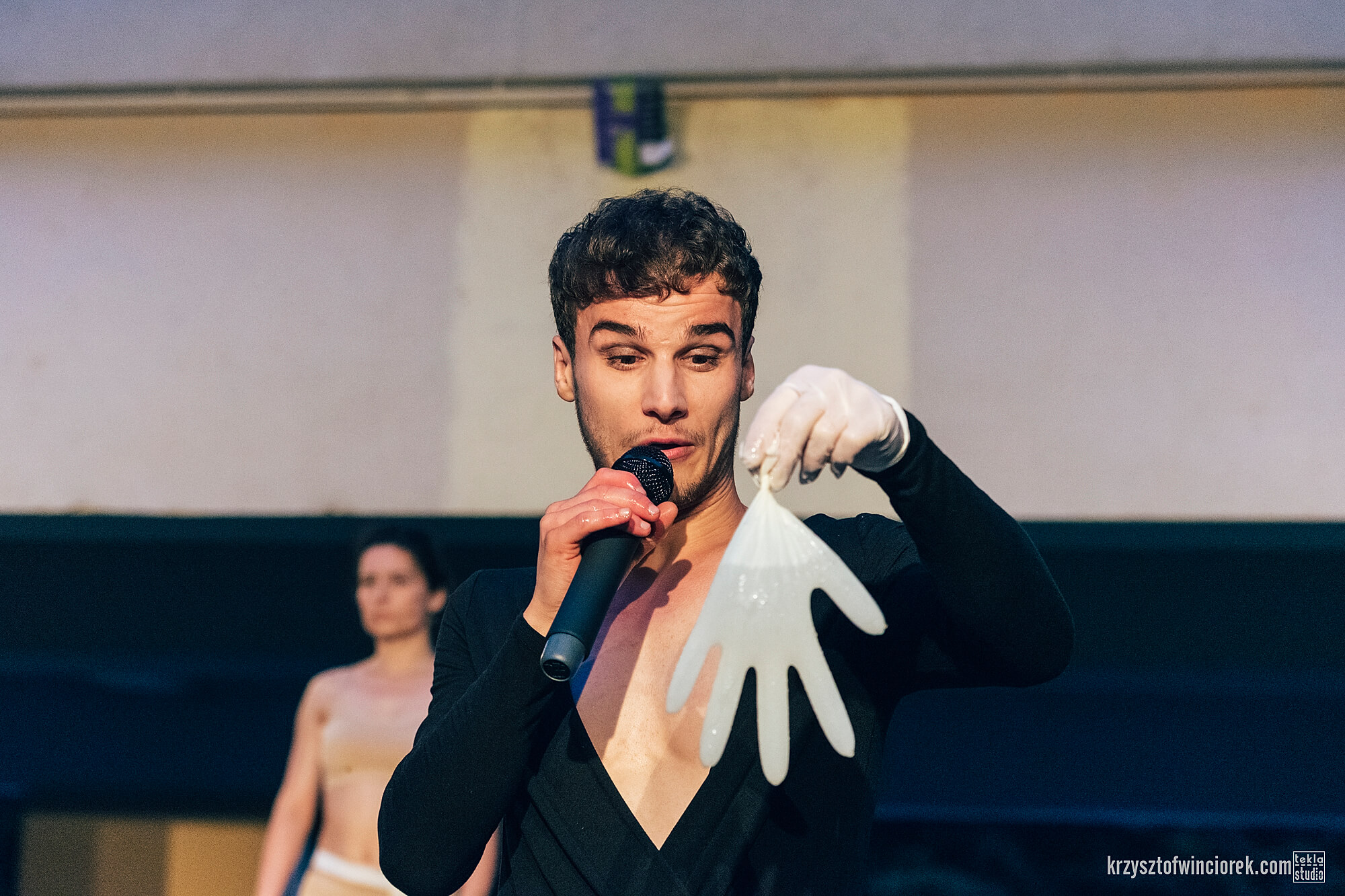 Zdjęcie z festiwalu Pociąg do miasta. Aktor w jednej ręce trzyma mikrofon, w drugiej białą lateksową rękawiczkę wypełnioną wodą.