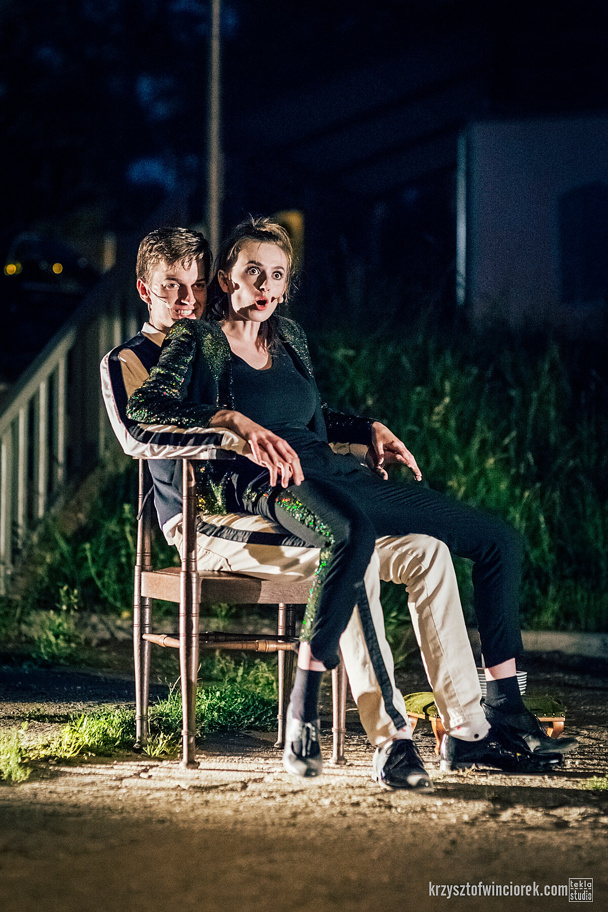 scena nocna, na zdjęciu dwójka aktorów, mężczyzna szczerząc się siedzi na drewnianym krześle, na jego kolanach siedzi aktorka w zielonym stroju